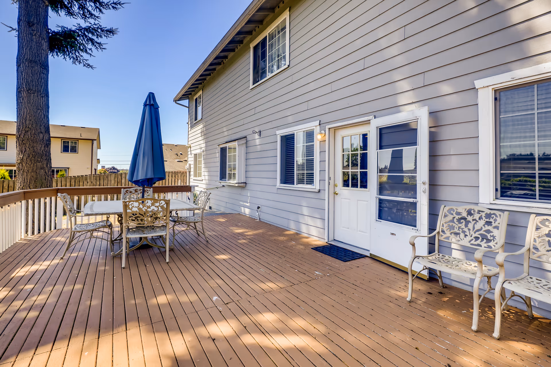 Outdoor wooden deck attached to a building with a white door and windows. The deck has a metal table with four matching chairs and a closed blue umbrella in the center. Two additional metal chairs are placed near the door. A tall tree and neighboring buildings are visible in the background under a clear blue sky.