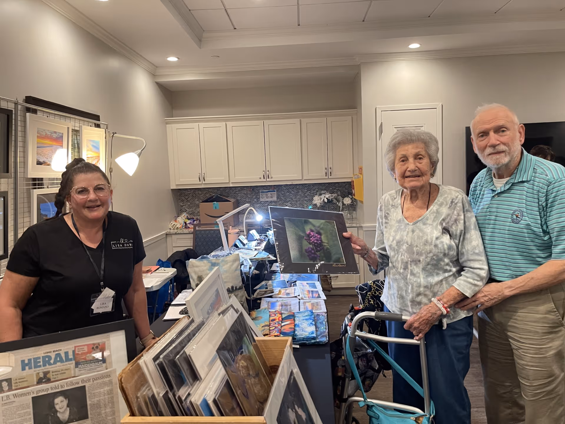 An elderly woman using a walker and an elderly man stand together inside a room, smiling at the camera. A woman wearing glasses and a black shirt with a name tag stands behind a table displaying various framed photographs and artwork. The room has white cabinets and soft lighting.