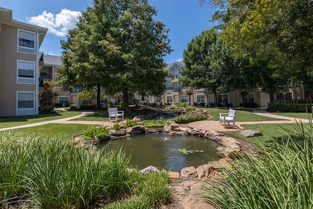 A landscaped outdoor courtyard at Brookdale First Colony featuring a small pond with rocks around it, green grass, trees, and two white benches facing the pond. Surrounding the courtyard are multi-story residential buildings under a partly cloudy blue sky.