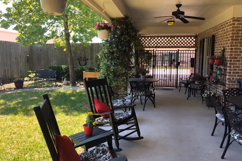 Covered patio area with black rocking chairs and small tables, decorated with red cushions and potted plants. The patio overlooks a grassy yard with a tree, a bench, and a wooden fence. Hanging flower pots and a ceiling fan are visible under the patio roof.
