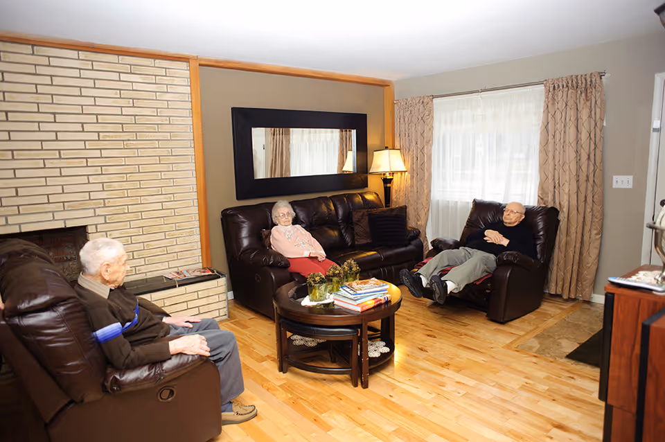 A cozy living room with three elderly people sitting on dark brown leather furniture. There is a brick fireplace on the left, a large rectangular mirror on the wall, a round wooden coffee table with books and decorative items, and a floor lamp next to a window with beige curtains.