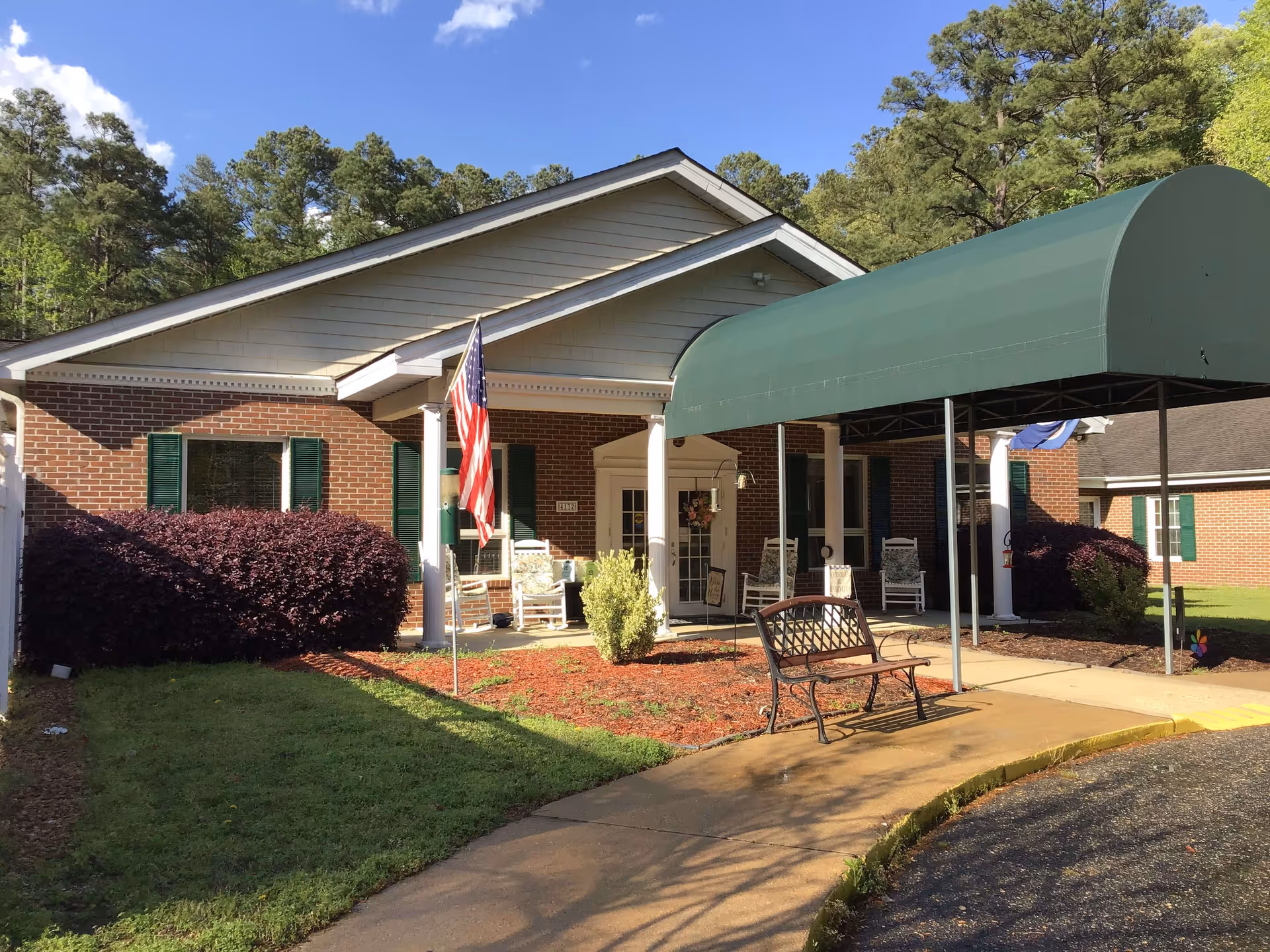 Exterior view of a single-story brick building with green shutters and a covered entrance canopy. There are rocking chairs on the porch, an American flag on a pole, a bench near the sidewalk, and well-maintained landscaping with bushes and grass. Trees and a blue sky with some clouds are visible in the background.