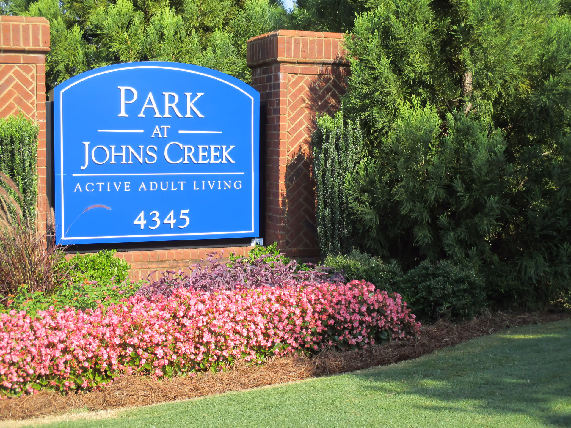 A blue sign with white text that reads 'Park at Johns Creek Active Adult Living 4345' set against a backdrop of green bushes and pink flowers, with brick pillars on either side.