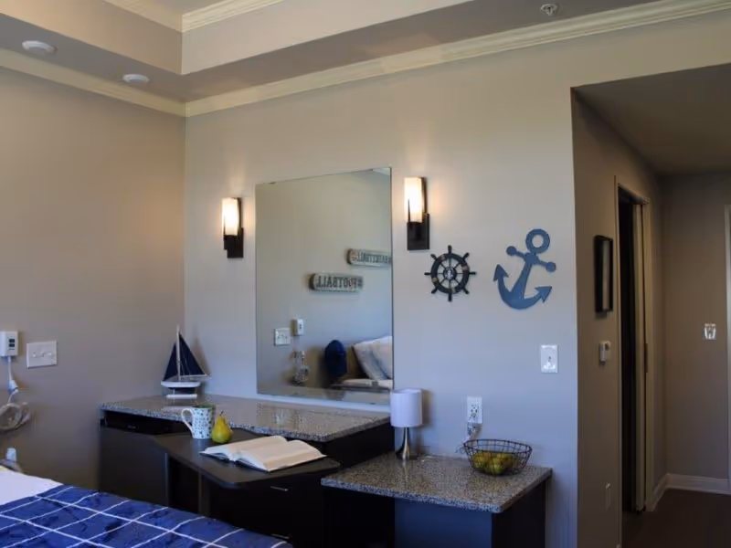 A senior living facility bedroom featuring a bed with a blue checkered blanket, a granite countertop with a small sailboat decoration, a mug, a pear, and an open book. On the wall above the countertop is a large mirror flanked by two wall sconces, and nautical-themed decorations including a ship's wheel and an anchor. A hallway is visible to the right.