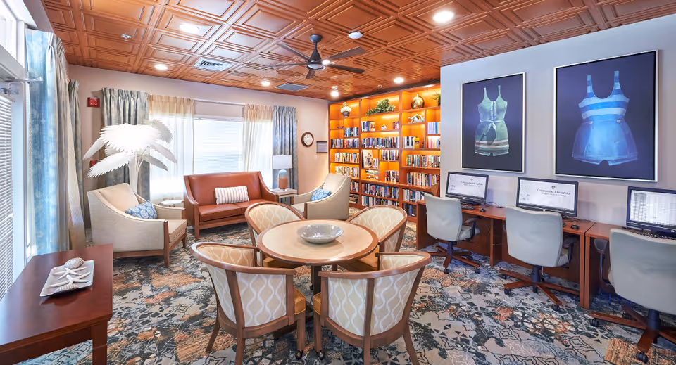 Bright communal lounge with a round table and chairs, sofas, bookshelves, and computer workstations.