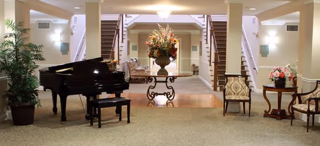 A spacious and elegant interior common area featuring a black grand piano with a matching bench on the left, a large floral arrangement on a decorative table in the center, and two sets of stairs leading upward on either side. There are upholstered chairs and small tables with flower arrangements on the right side, soft lighting fixtures on the walls, and a large potted plant on the far left.