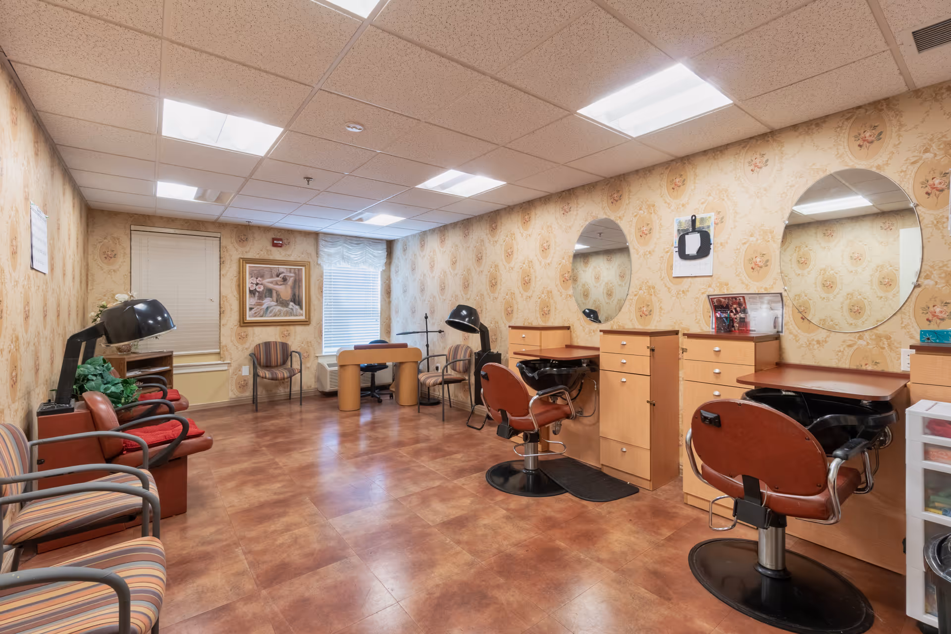 Interior view of a salon area in a senior living facility with two styling chairs in front of mirrors, a hair washing station, several waiting chairs, a small table with two chairs near the window, and floral wallpaper on the walls.