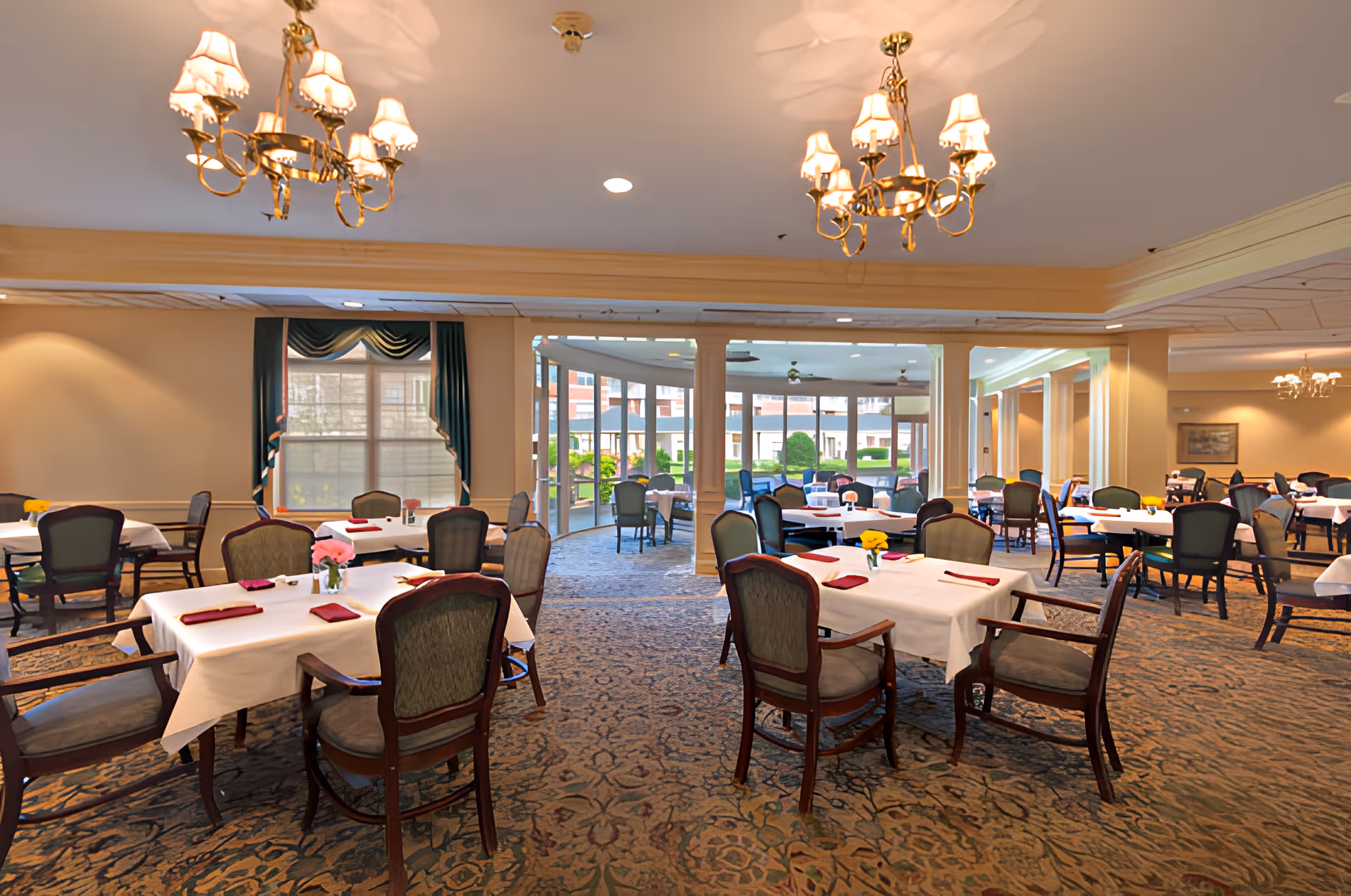 Spacious dining room with multiple tables set with white tablecloths and chairs under chandeliers, opening to a bright sunroom.