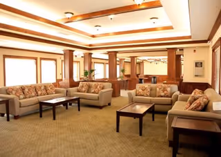 A spacious living room area with beige sofas and floral patterned cushions arranged around dark wooden coffee tables. The room features large windows with white blinds, wooden columns, and a coffered ceiling with recessed lighting.