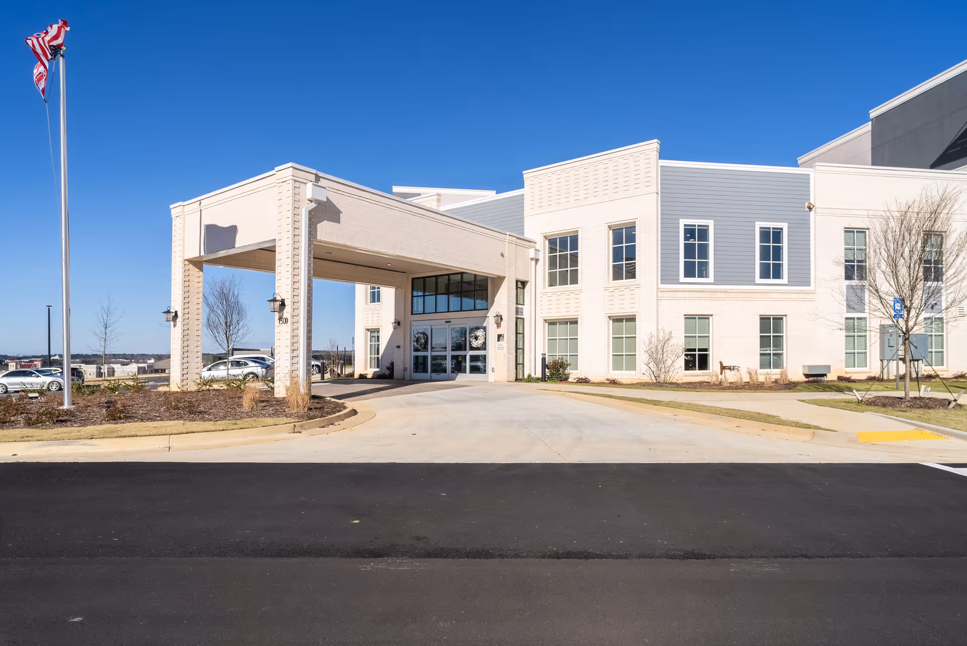 Front exterior view of a modern senior living facility building with a covered entrance, large windows, and an American flag on a flagpole to the left. The sky is clear and blue.