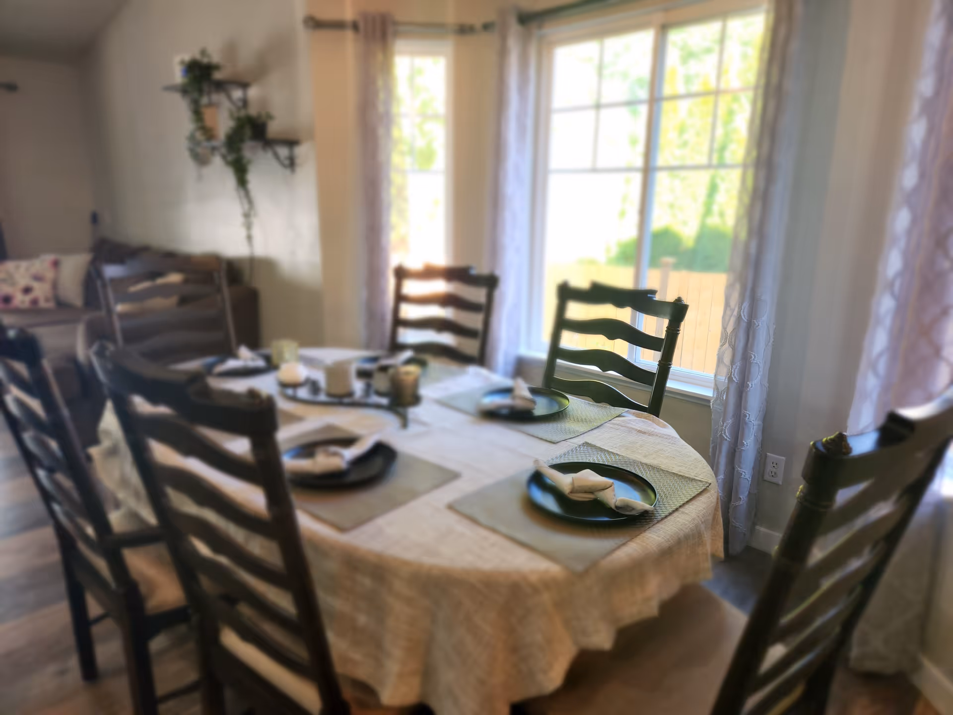 Dining room with a table set with plates and napkins surrounded by wooden ladder-back chairs near a large sunny window.