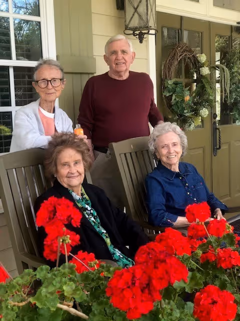 Four elderly individuals, two men and two women, are gathered on a porch with green siding and a door decorated with a wreath. Two women are seated in wooden rocking chairs surrounded by vibrant red flowers, while the other two stand behind them. The setting appears warm and inviting with natural light.