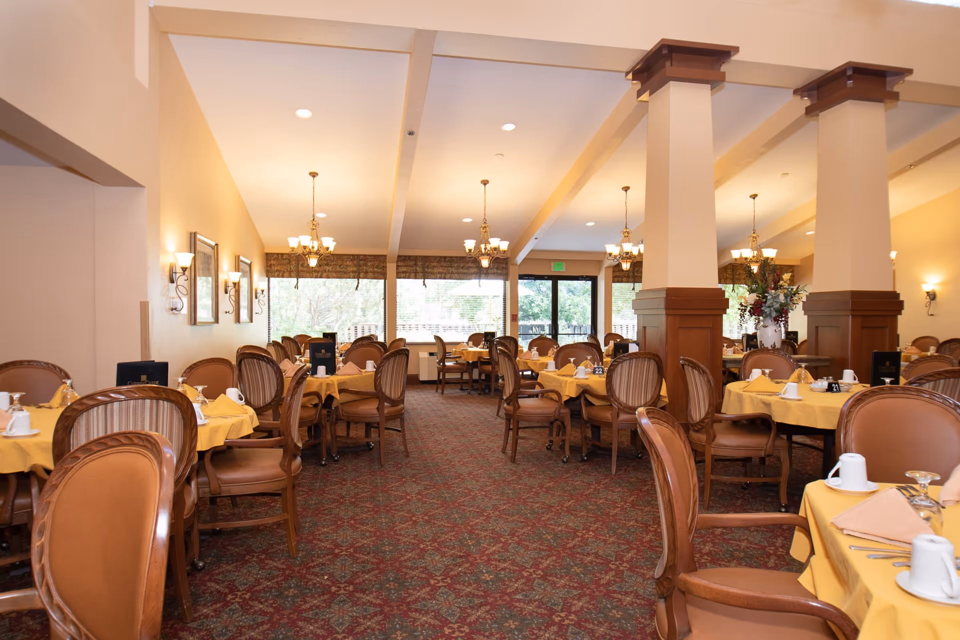 A spacious dining room with multiple round tables covered with yellow tablecloths, each set with white cups, glasses, and napkins. The room features brown upholstered chairs, carpeted flooring with a patterned design, beige walls, and large windows letting in natural light. There are decorative wall sconces and chandeliers hanging from the ceiling, and large wooden columns in the center of the room.
