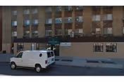 Street view of a multi-story brick building with several windows, some with air conditioning units. A white van is parked on the street in front of the building. The entrance has a green awning with text, and there are signs on the building's facade.
