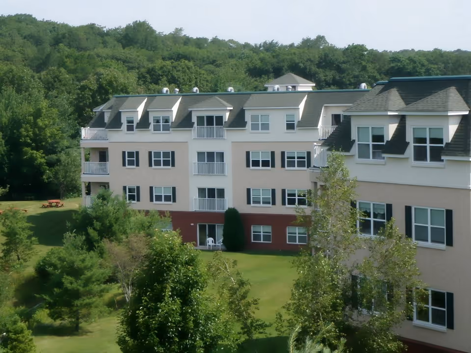 Exterior view of a multi-story senior living facility building surrounded by green trees and grass, with a picnic table visible on the lawn.