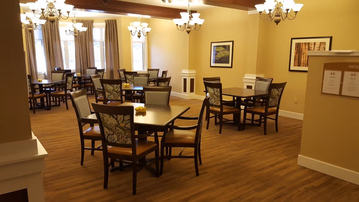 A dining room with several dark wood tables and chairs with patterned backs. The room has wood flooring, beige walls, and multiple chandeliers hanging from the ceiling. There are framed pictures on the walls and windows with curtains letting in natural light.