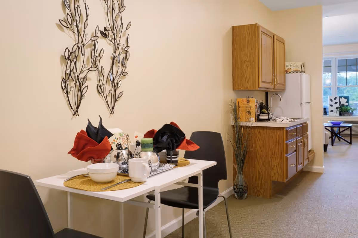 A small dining area with a white table set for two, featuring black and red folded napkins, a bowl, a mug, and utensils. Behind the table is a wall decorated with metal leaf art. To the right, there is a kitchenette with wooden cabinets, a sink, and a white refrigerator. In the background, a living room area with a window and a coffee table is partially visible.