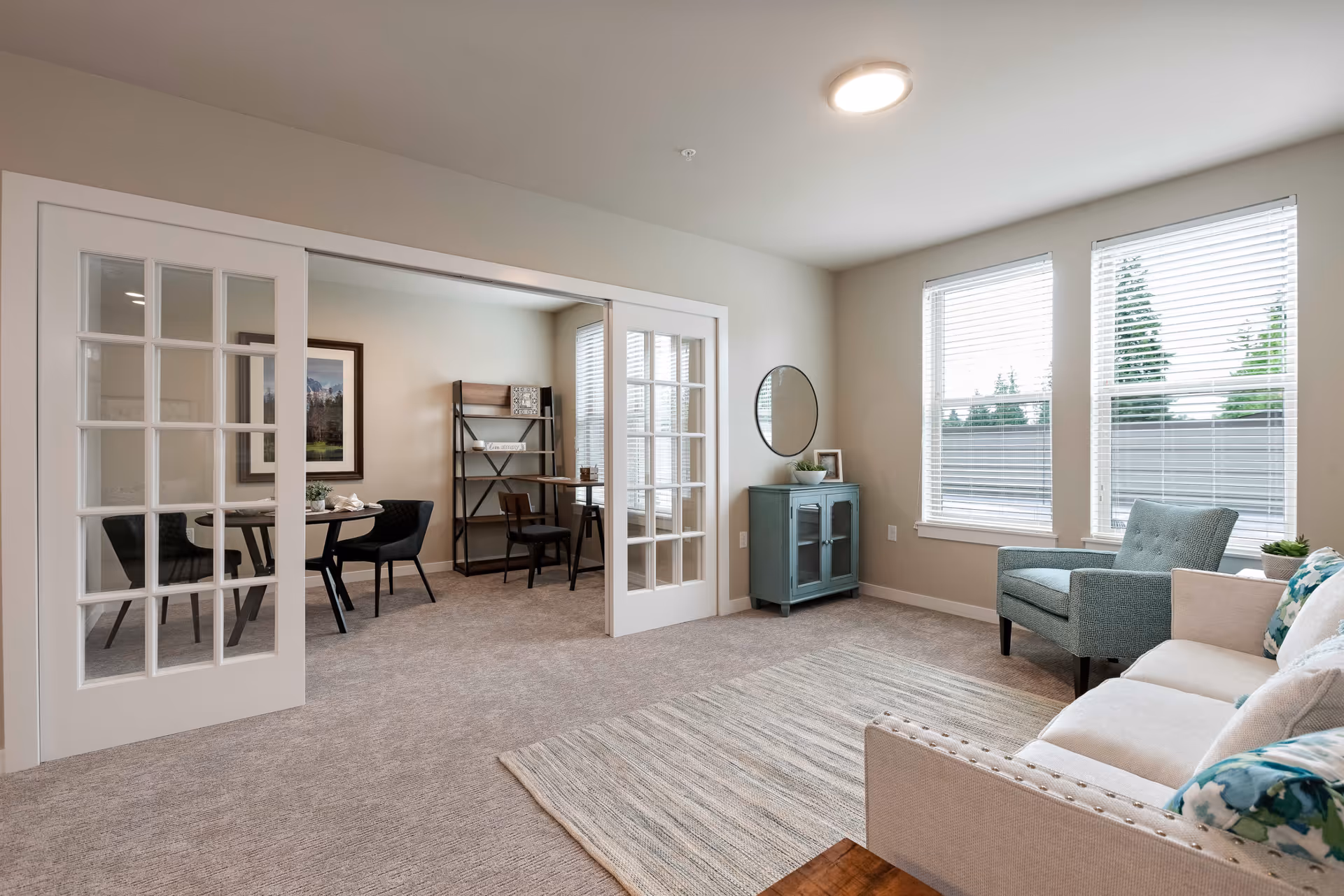 A bright and cozy living room with a beige sofa adorned with floral cushions, a light blue armchair, and a striped area rug on carpeted floor. Two large windows with white blinds let in natural light. A small teal cabinet with a round mirror above it is placed against the wall. Adjacent to the living room is a room separated by white sliding glass doors, featuring a round table with black chairs and a wooden desk with a chair and shelving unit.