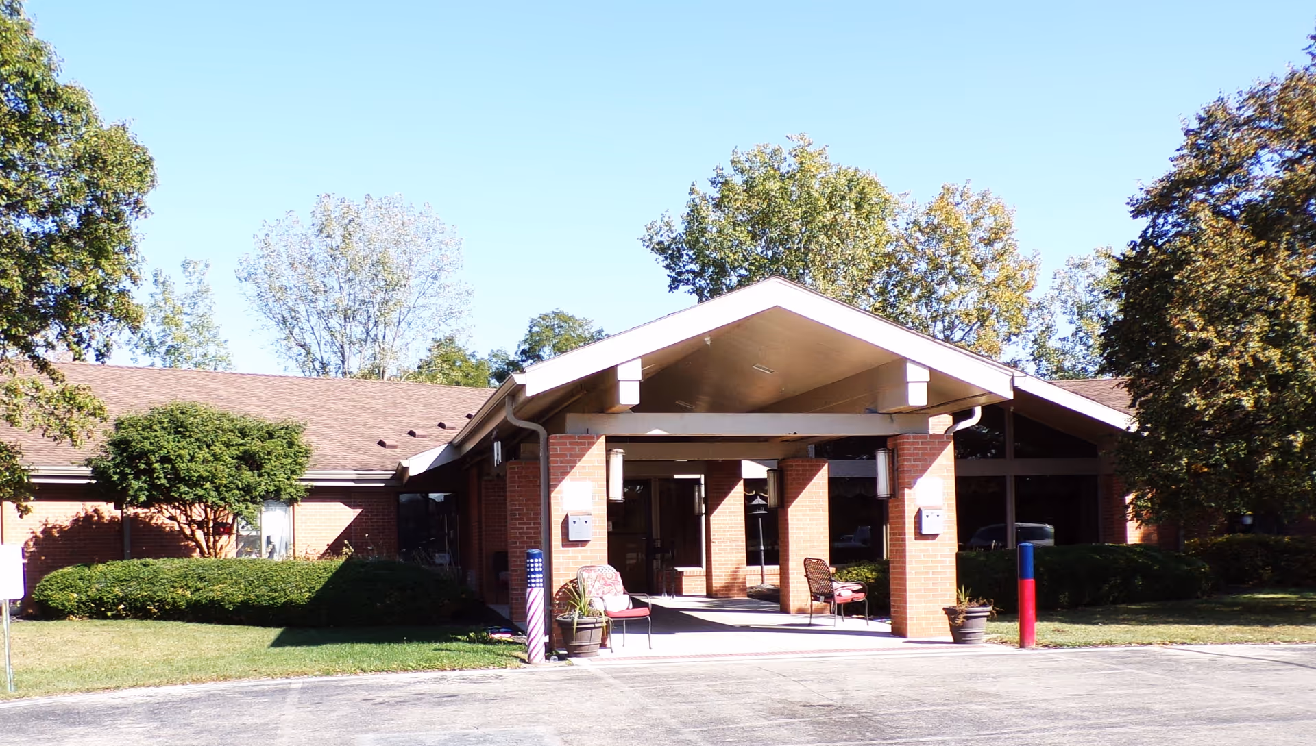 Front exterior view of a single-story brick building with a covered entrance, two chairs, potted plants, and surrounding trees and bushes under a clear blue sky.