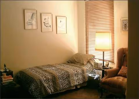 A cozy bedroom with a single bed, nightstand, lamp, and armchair by a window.