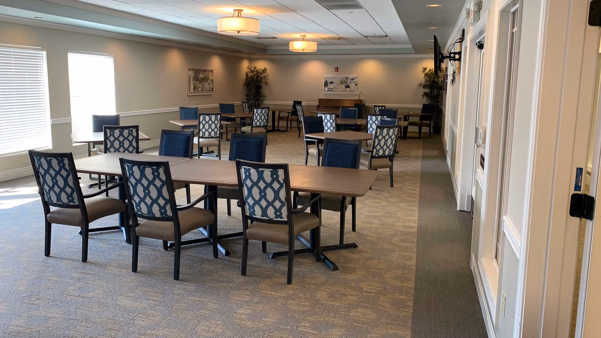 A spacious dining room with multiple rectangular tables and chairs arranged neatly. The chairs have patterned backs and cushioned seats. The room is well-lit with natural light coming from large windows on the left and ceiling lights. There are plants and framed pictures on the walls, and a piano is visible at the far end of the room.