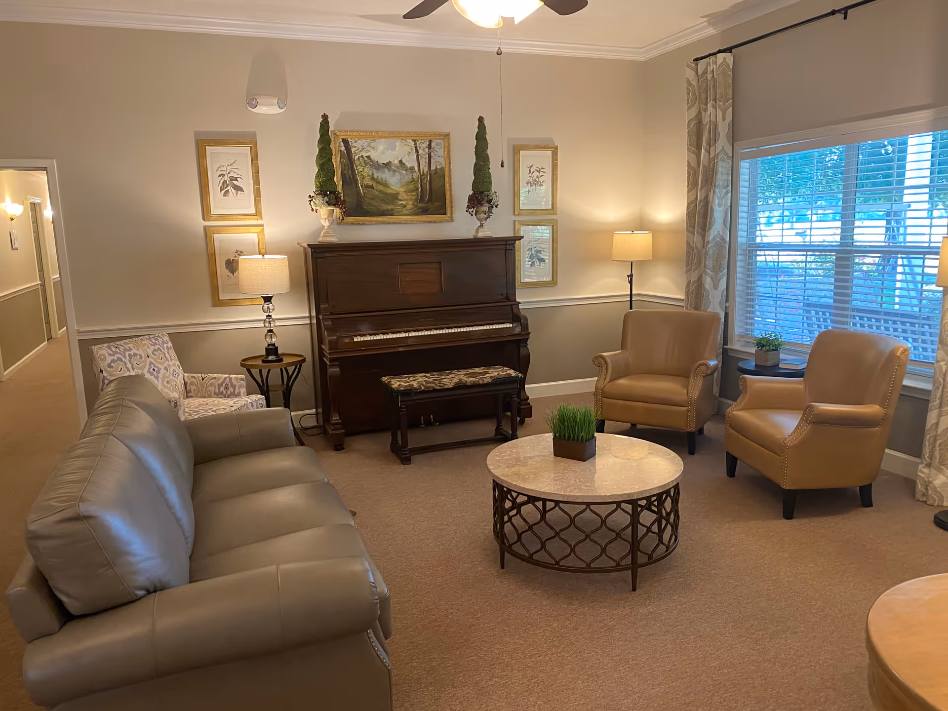 A cozy living room with a beige carpet, a gray leather sofa, two tan armchairs, and a patterned armchair. There is a round coffee table with a small plant in the center. A dark wooden upright piano with a cushioned bench is against the wall, flanked by two tall decorative plants and framed artwork. The room is softly lit by table lamps and a floor lamp, with a large window covered by patterned curtains letting in natural light.