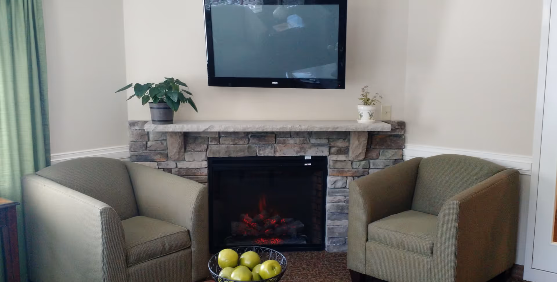 A cozy seating area with two green armchairs facing a stone fireplace with a small fire burning. Above the fireplace is a mounted flat-screen TV. A small table in front of the fireplace holds a bowl of green apples. There are two potted plants on the fireplace mantel, and green curtains are visible on the left side.