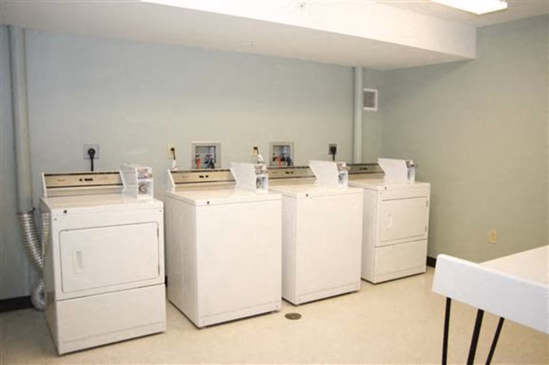 A laundry room with four white washing machines or dryers lined up against a light green wall. There is a white table with black legs partially visible on the right side of the image.