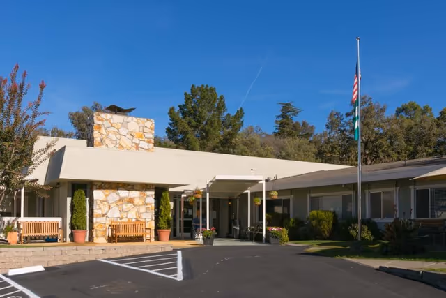 Front exterior of a single-story senior living facility showing a covered entrance with a stone chimney, benches, flagpole, and parking area.