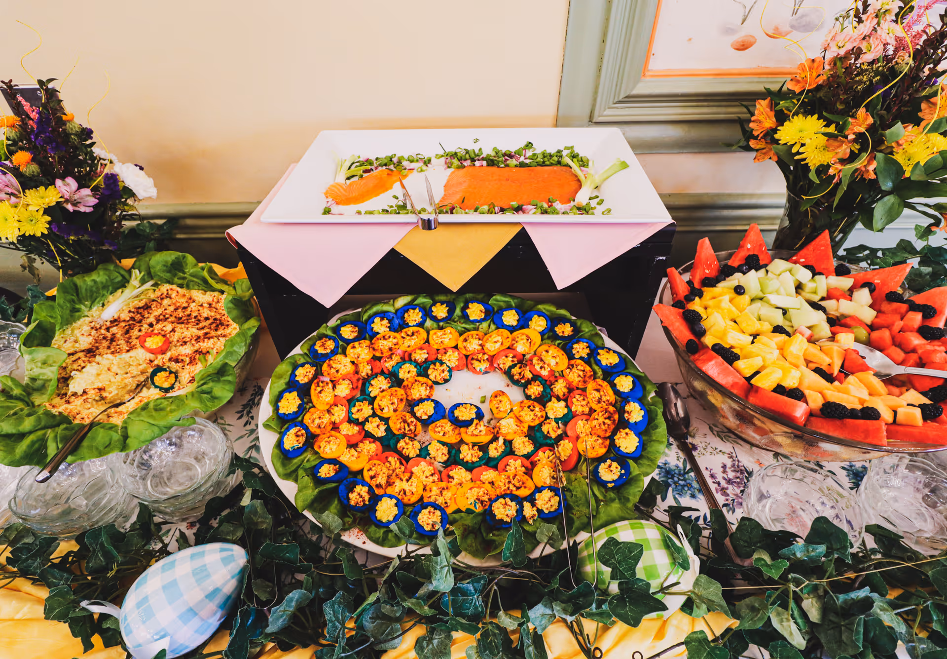 A buffet table with a variety of foods including a large bowl of mixed fruit, a platter of deviled eggs arranged in a spiral pattern, a dish of smoked salmon garnished with greens, and a bowl of salad. The table is decorated with green ivy and colorful flowers in vases.