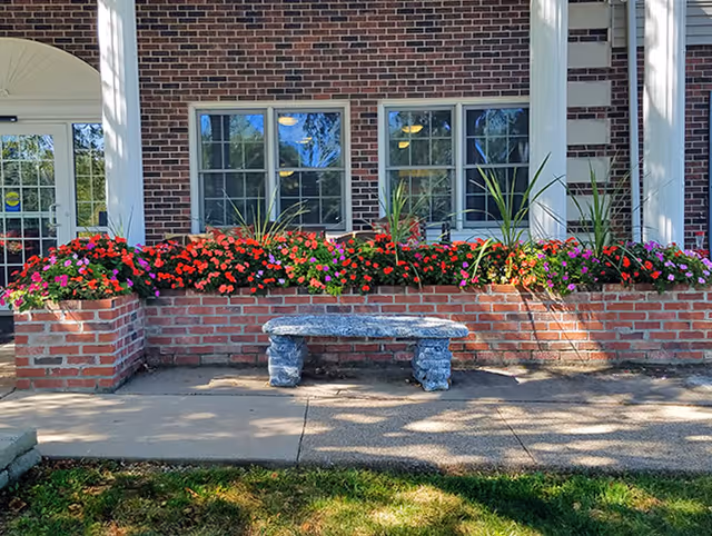 Brick building front with a raised planter full of colorful flowers and a stone bench on the sidewalk.