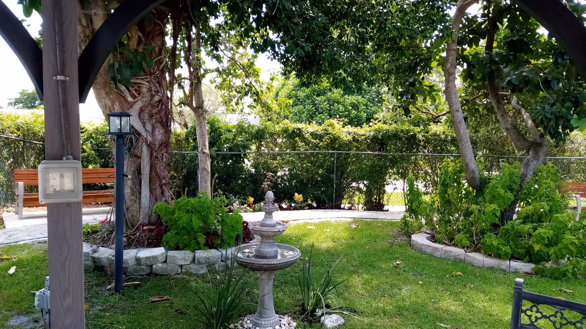 A peaceful outdoor garden area with green grass, various plants, and trees. There is a small stone fountain in the center, a wooden bench in the background, and a black metal lamp post near a large tree. The area is partially shaded by a wooden pergola structure.