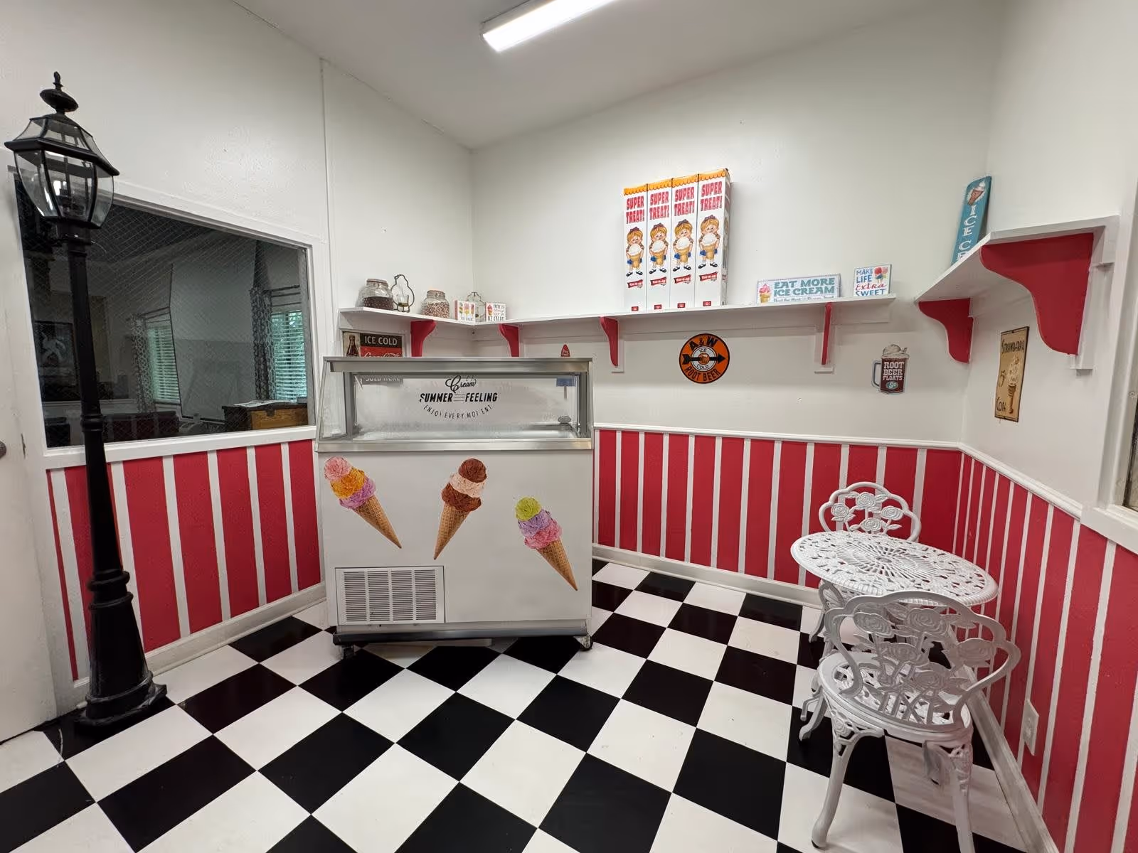 A small ice cream parlor area with a black and white checkered floor, pink and white striped walls, and white decorative metal table and chairs. There is an ice cream freezer with images of ice cream cones on it, a black street lamp, and shelves with vintage-style signs and boxes of Super Treat cereal.