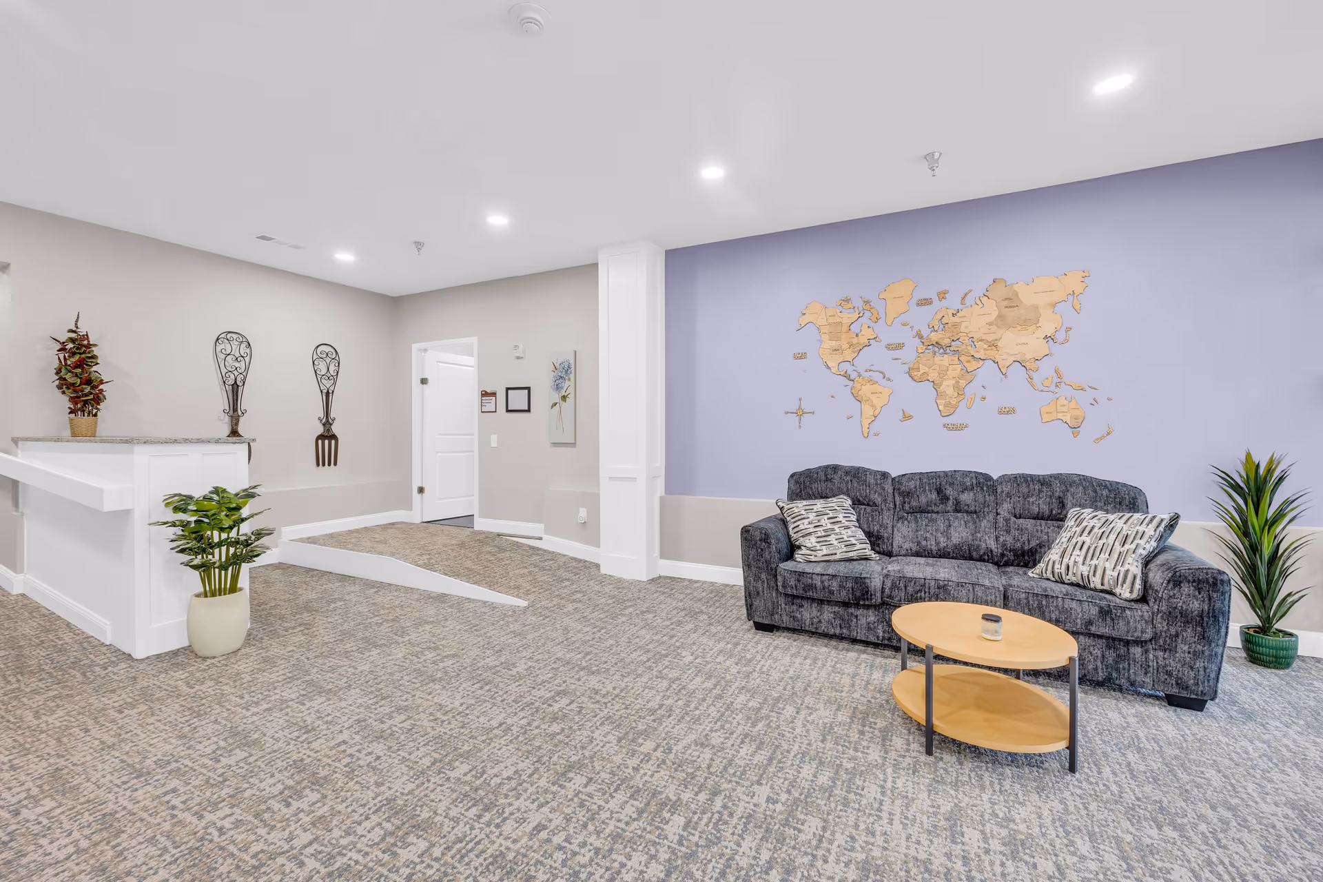 A bright and spacious senior living common area with a gray couch adorned with patterned pillows, a small wooden coffee table, and a decorative wooden world map mounted on a lavender accent wall. The room has light-colored walls, recessed ceiling lights, carpeted flooring, and several potted plants. There is also a small ramp leading to a doorway and decorative wall hangings shaped like a fork and spoon.