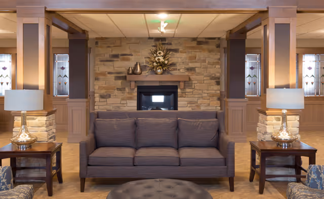 A cozy living room area with a gray three-seater sofa centered in front of a stone fireplace. On either side of the sofa are wooden side tables with glass lamps. The room features wooden pillars and stained glass windows in the background.