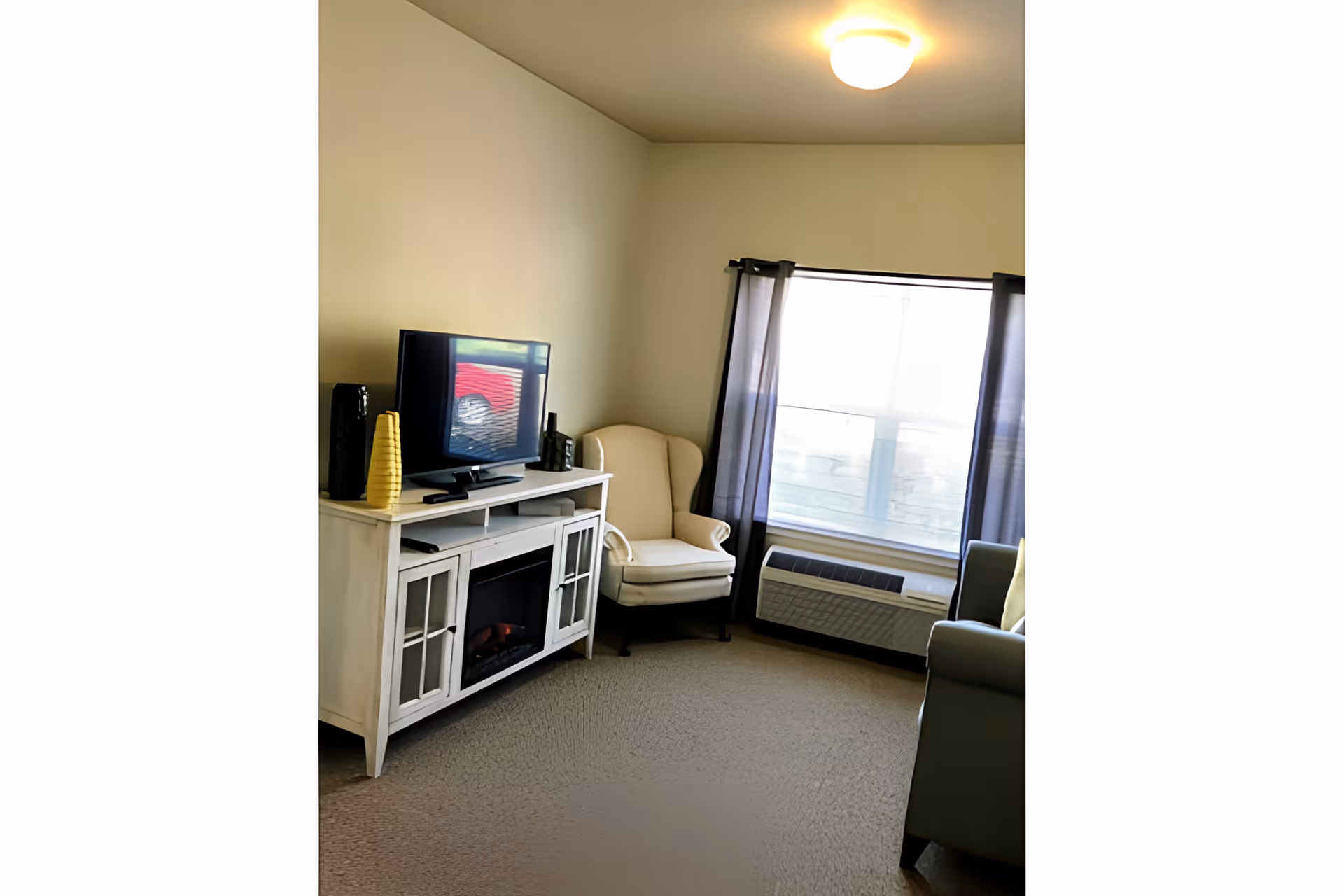 A cozy living room area with a white TV stand holding a flat-screen television and decorative vases. Next to the TV stand is a beige armchair, and a window with sheer black curtains lets in natural light. A green armchair is partially visible on the right side, and a ceiling light fixture illuminates the room.