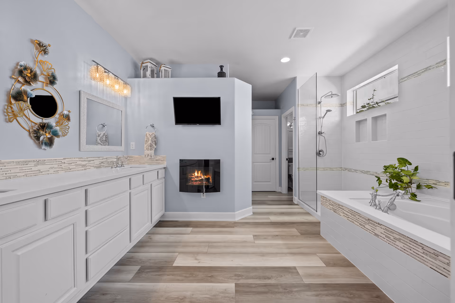 Spacious modern bathroom with a long white double vanity, built-in bathtub, glass shower, wall-mounted TV and a fireplace.