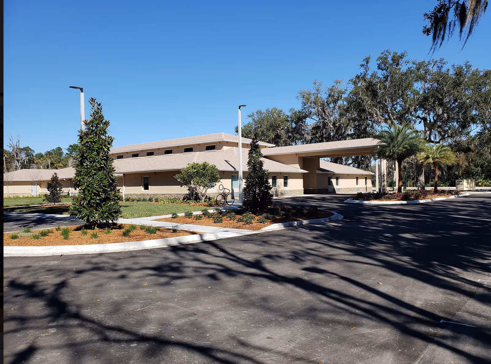 Exterior view of Ormond Manor facility showing a single-story beige building with a covered entrance, surrounded by landscaped greenery, trees, and a clear blue sky.
