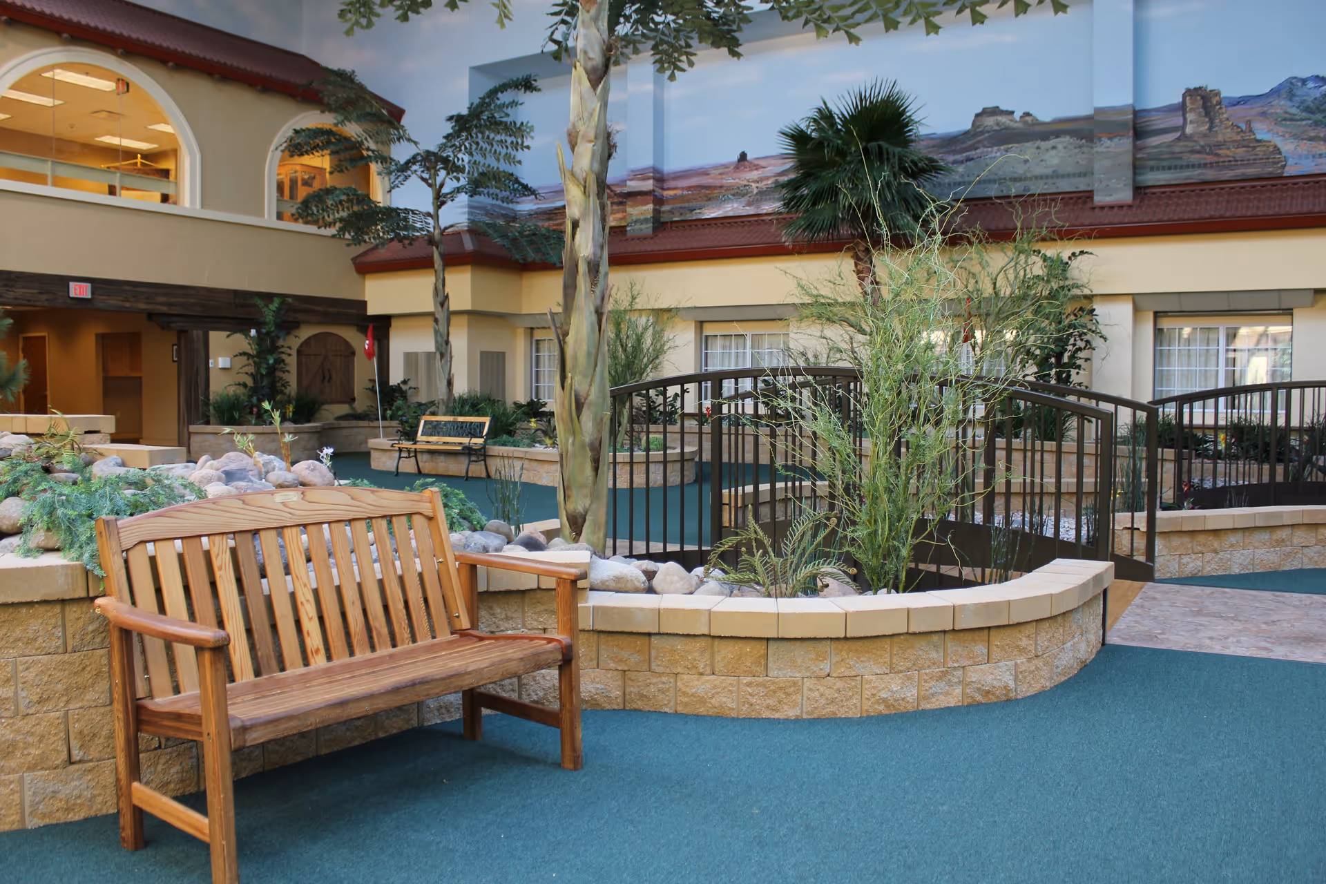 Indoor courtyard area with a wooden bench, plants, small trees, and a decorative stone planter. There is a small bridge over a recessed area and windows of the facility visible in the background. The walls feature a mural of a desert landscape.