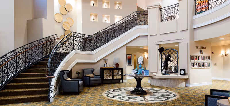 Spacious and elegant interior of a senior living facility featuring a grand staircase with ornate black railings, comfortable seating areas with armchairs, decorative wall niches with small sculptures, a round table with decorative items in the center, and a patterned carpet floor.
