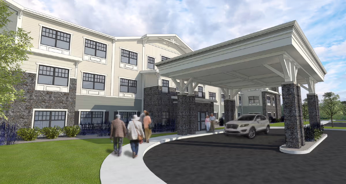 Exterior view of a senior living facility named Traditions of Columbus showing a covered entrance with stone pillars, a driveway with a parked car, and several people walking towards the building. The building is three stories tall with large windows and a combination of stone and light-colored siding.