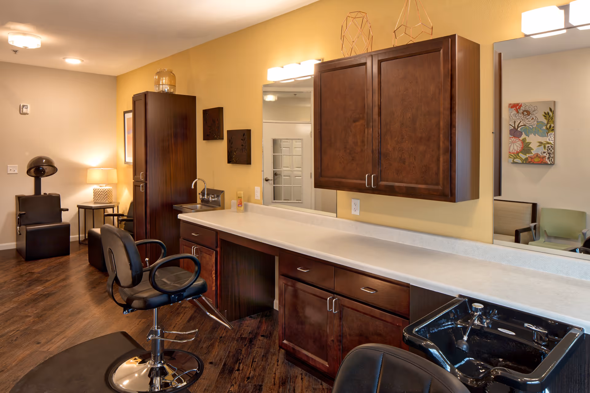 Interior of a hair salon area in an assisted living facility featuring salon chairs, a hair washing station with a black sink, wooden cabinets, a long countertop, and warm lighting. The room has wooden flooring and a cozy ambiance with a lamp and artwork on the walls.