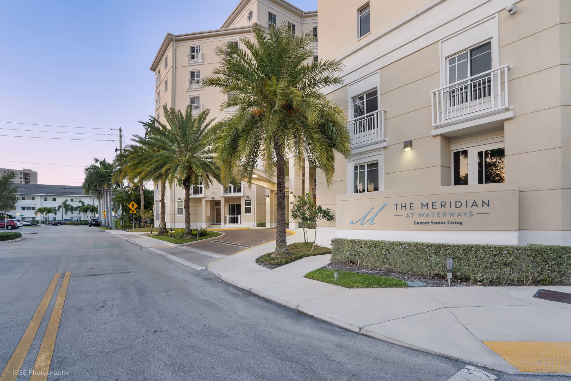 Exterior view of The Meridian at Waterways senior living facility showing a multi-story beige building with palm trees along the driveway and a sign that reads 'The Meridian at Waterways Luxury Senior Living'.