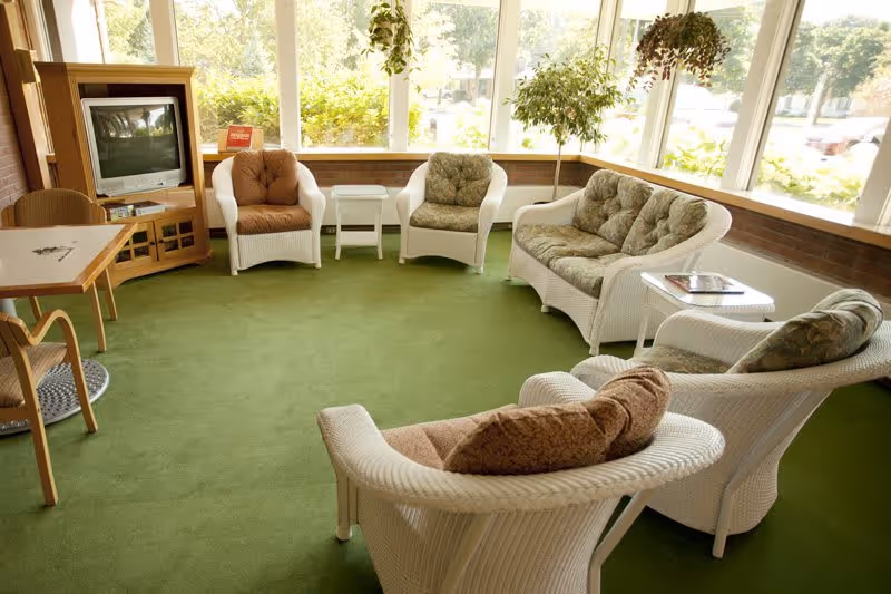 A bright living room area with green carpet, white wicker furniture including a loveseat, four armchairs with cushions, a small white side table, and a wooden TV stand with an older model television. Large windows let in natural light and show greenery outside.