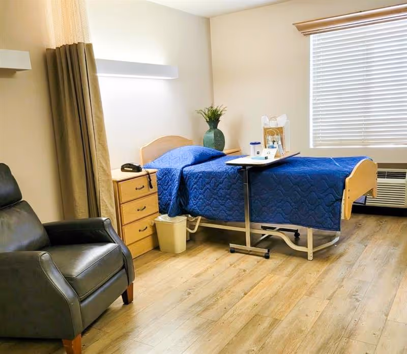 A neatly arranged room with a single hospital-style bed covered with a blue quilt, a wooden nightstand with a phone and a green vase with flowers, a black leather armchair, a window with blinds, and a wooden floor.