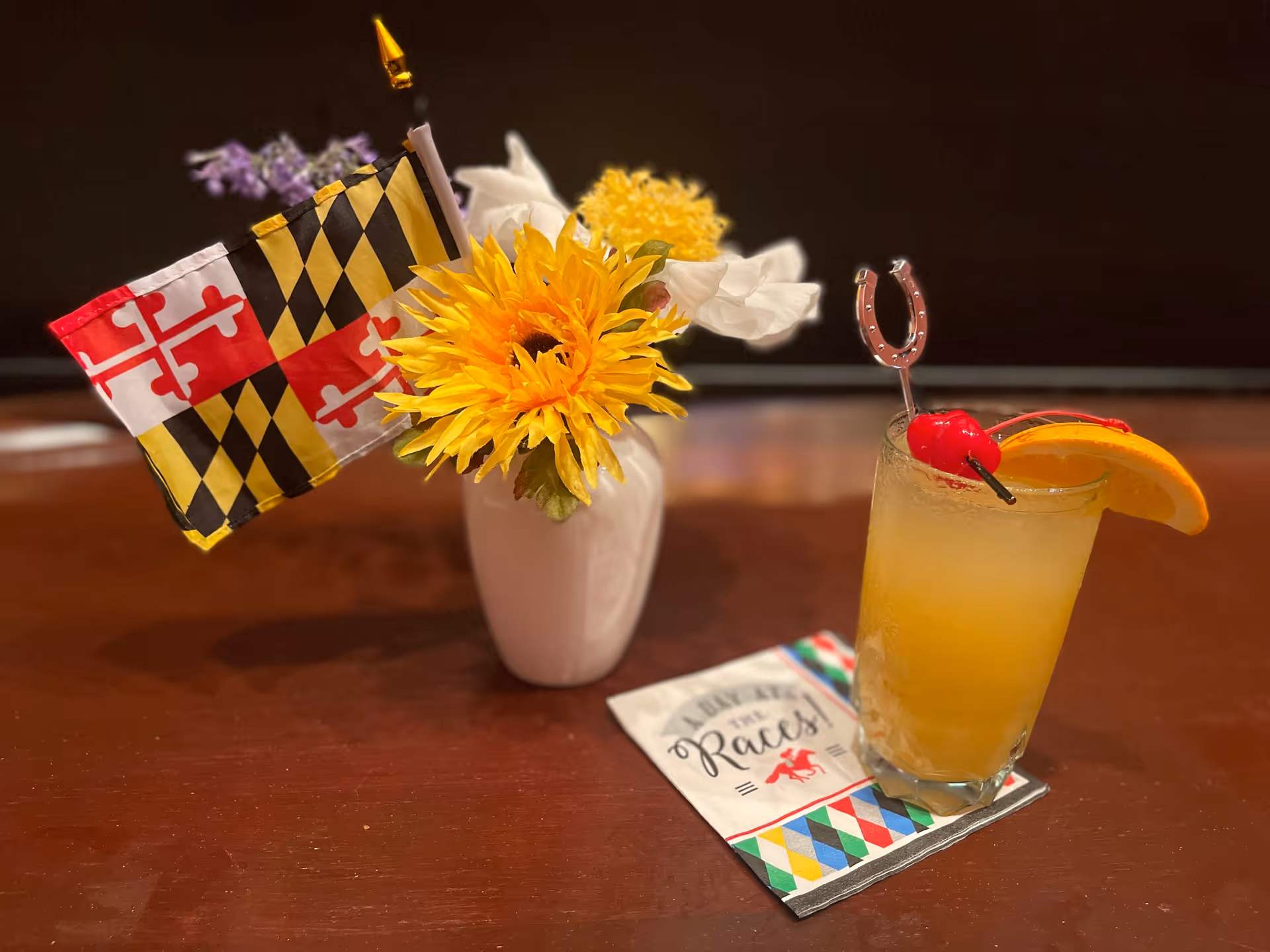 A close-up of a table with a small white vase holding a bouquet of flowers including a sunflower and a small Maryland state flag. Next to the vase is a glass of orange-colored drink garnished with a cherry, an orange slice, and a horseshoe-shaped stirrer. A colorful napkin with the text 'I bet at the Races!' is placed under the glass.