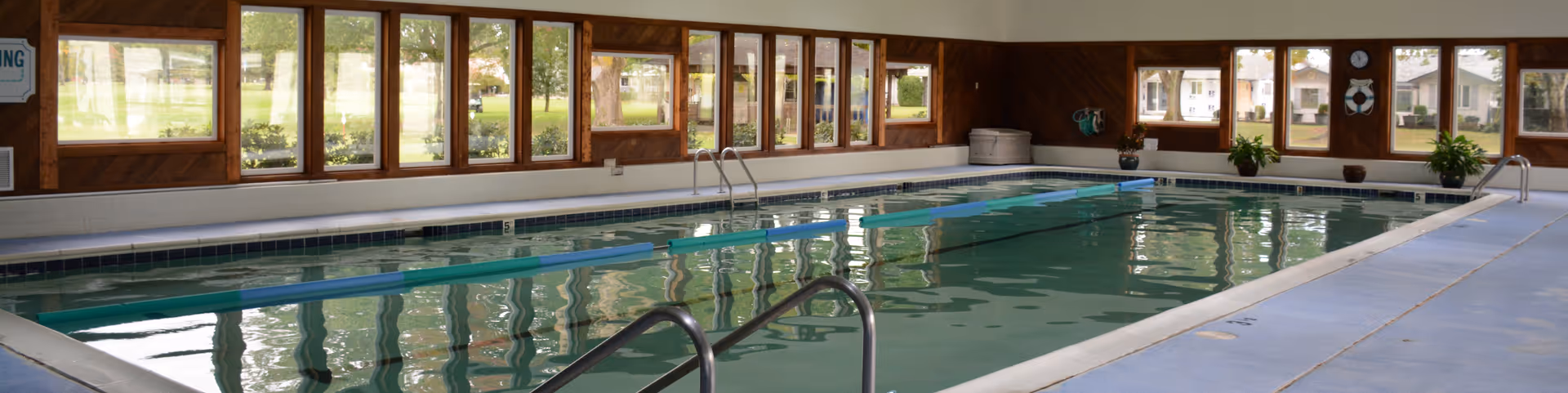 Indoor swimming pool with handrails and large windows along the walls allowing natural light to enter. The pool area has a blue floor and wooden paneling on the walls, with some potted plants and safety equipment visible.