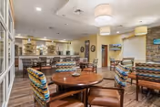 Communal dining room with round wood tables, patterned upholstered chairs, a bar seating area and warm overhead lighting.
