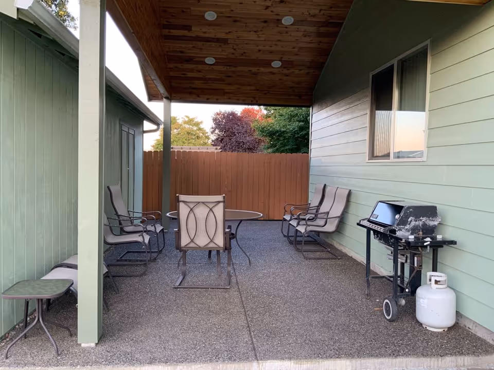 Covered outdoor patio with several metal framed chairs around a round table and a propane grill next to a green house wall.