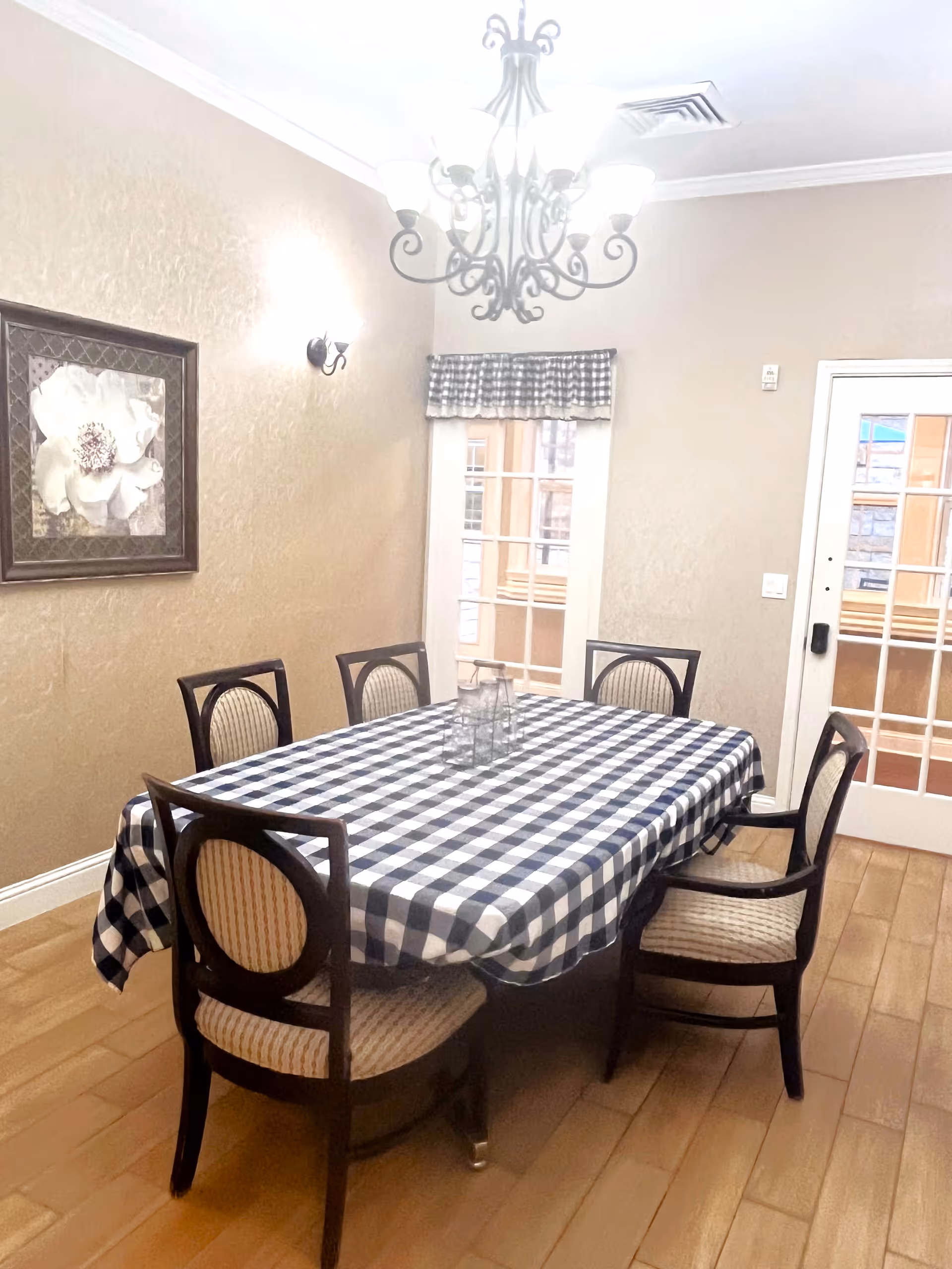 A dining room with a rectangular table covered with a black and white checkered tablecloth. Six wooden chairs with cushioned seats surround the table. A chandelier with multiple lights hangs above the table. There is a framed floral artwork on the beige wall to the left, and a window with a checkered valance and a glass door are visible on the far wall. The floor is wooden.